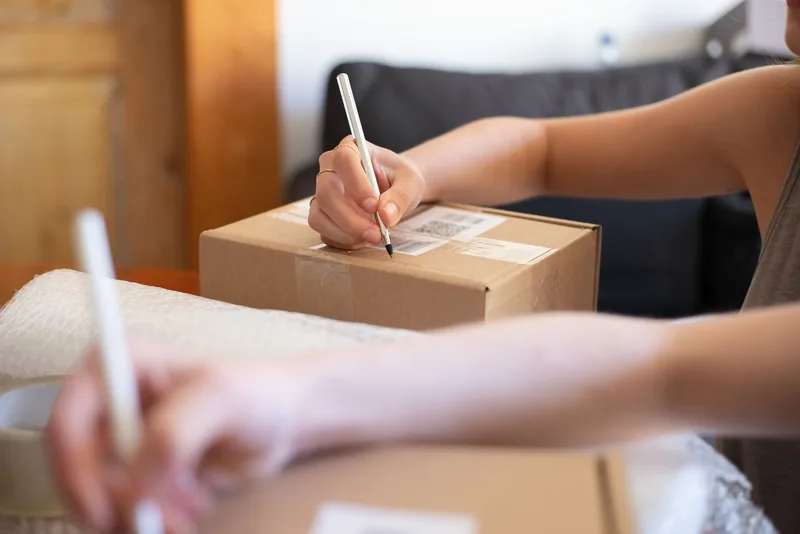 Close-up of hands labeling a cardboard box for shipping from a home ecommerce workspace